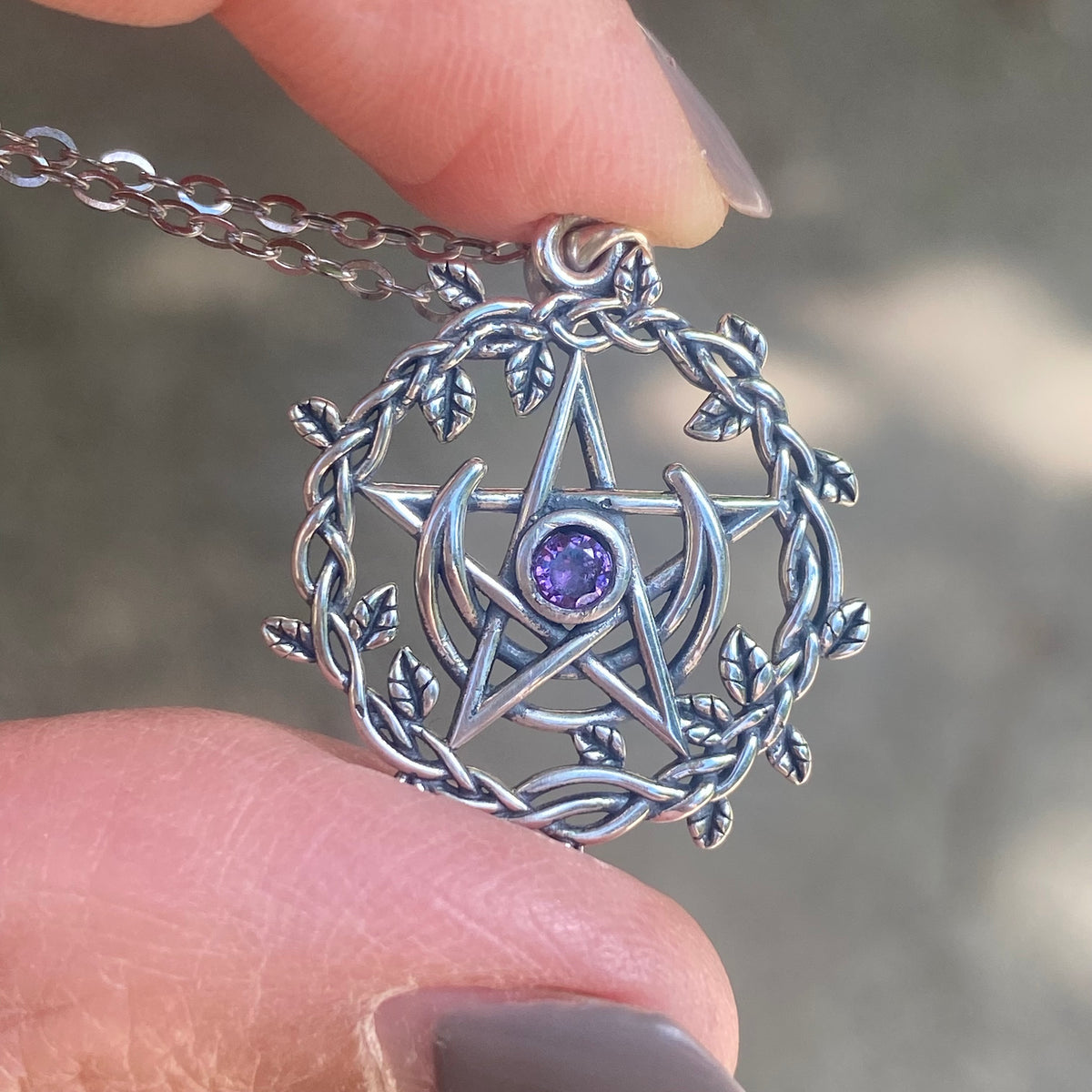 Sterling silver pentagram necklace with leaf wreath design and purple gemstone held between fingers